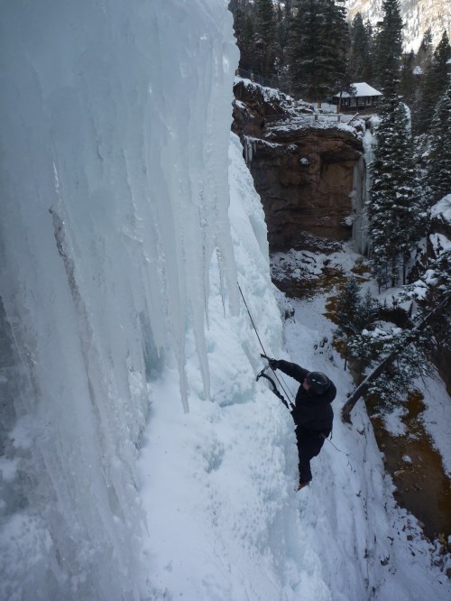 A climber ascends a frozen waterfall, surrounded by snow-covered trees and rocky cliffs.