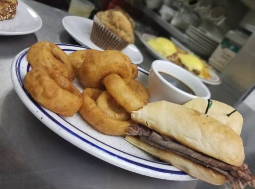 A plate with a sandwich filled with roast beef, onion rings, and a small cup of dipping sauce in the background.