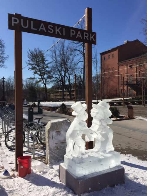 Ice sculpture of two figures holding hands in front of the Pulaski Park sign, surrounded by snow and trees.