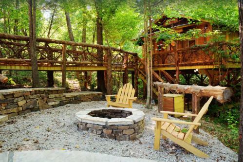 A cozy outdoor fire pit area with two chairs, surrounded by trees and a wooden structure in the background.