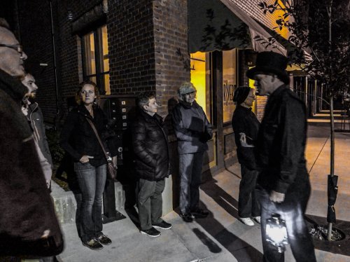 A group of people stands outside a building at night, some looking at a man holding a lantern.