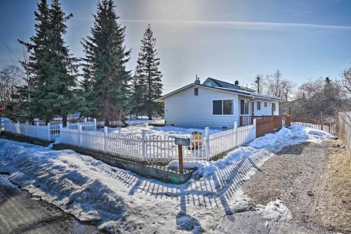 A white house with a fenced yard, surrounded by snow and tall trees, under a clear blue sky.