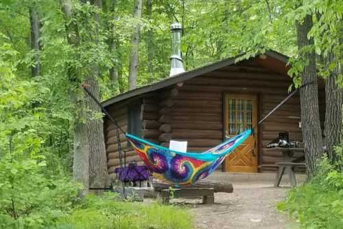 A colorful hammock hangs between trees in front of a rustic log cabin surrounded by lush greenery.