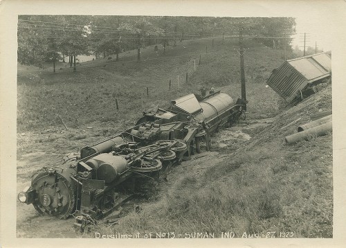 A vintage photo of a derailed steam locomotive in a rural area, with trees and a hillside in the background.