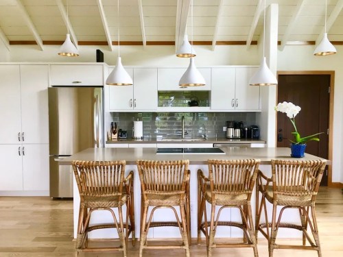 Modern kitchen with white cabinets, a large island, rattan bar stools, and pendant lights, featuring a potted orchid.