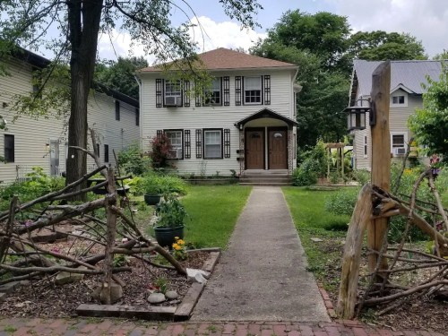 A charming two-story house with a front garden, surrounded by greenery and a rustic wooden gate.