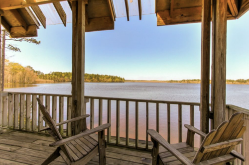 View from a wooden porch overlooking a calm lake surrounded by trees under a clear blue sky.