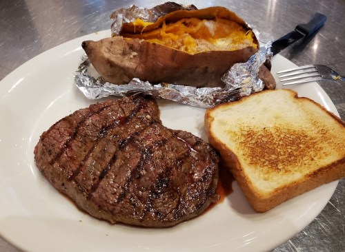 A plate with a grilled steak, a baked sweet potato wrapped in foil, and a slice of toasted bread.
