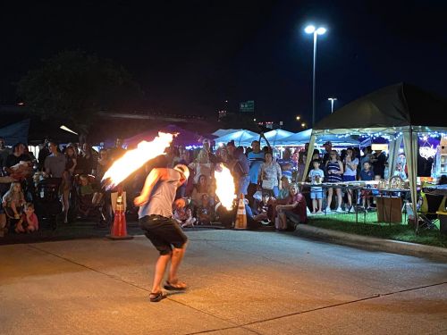 A fire performer spins flaming torches in front of a crowd at a night market, with tents and lights in the background.