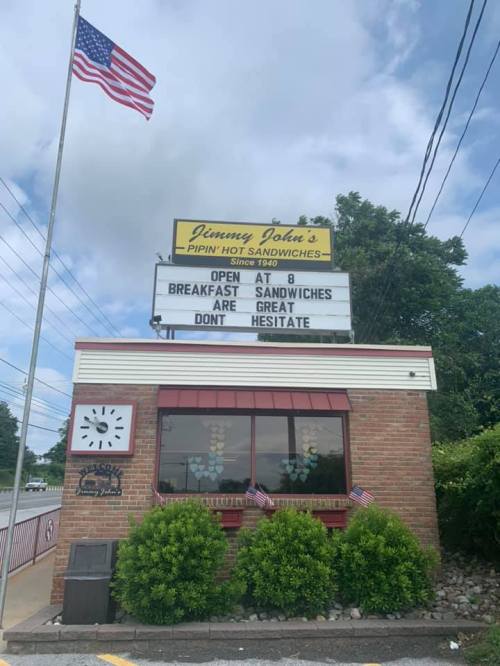 Sign for Jimmy John's with American flag, promoting breakfast sandwiches and encouraging customers not to hesitate.