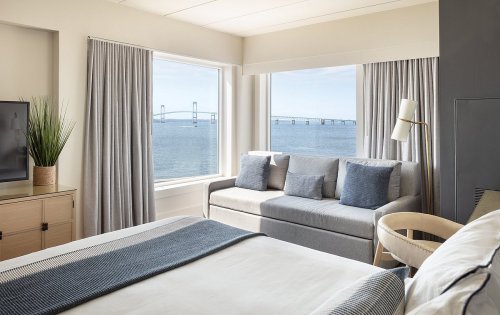 A modern hotel room with a bed, sofa, and large windows showcasing a view of the ocean and a bridge.