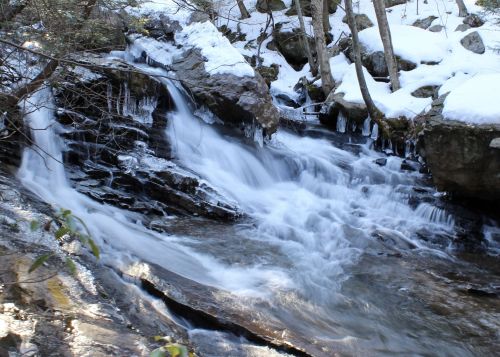 A serene waterfall cascades over rocks, surrounded by snow-covered trees and a tranquil winter landscape.