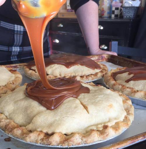 A person drizzling caramel sauce over unbaked pies on a tray, with a kitchen background.