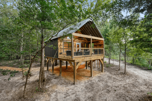 A wooden cabin on stilts surrounded by trees, featuring a large deck and open design in a natural setting.