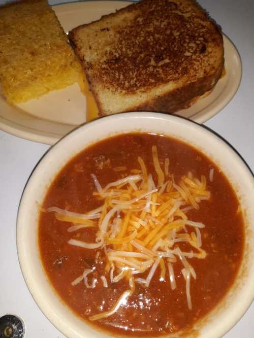 A bowl of chili topped with shredded cheese, accompanied by a plate of cornbread and toasted bread.