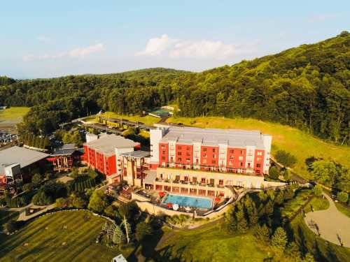 Aerial view of a red hotel surrounded by greenery, featuring a pool and landscaped gardens.