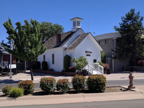 A small white historic building with a peaked roof, surrounded by greenery and a clear blue sky.