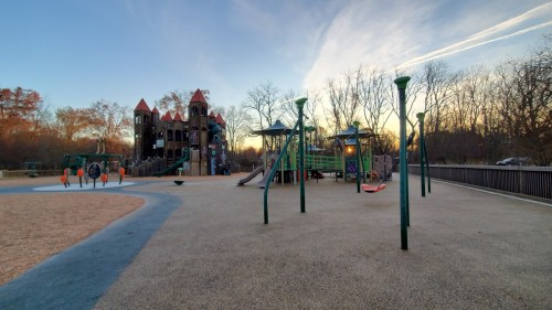 A playground with colorful equipment, including slides and swings, set against a sunset sky and bare trees.