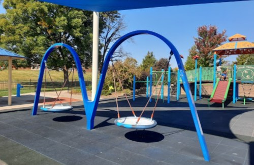 Two blue swings with circular seats in a colorful playground under a shaded structure, surrounded by trees.