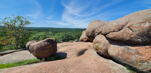 A scenic view of large boulders on a rocky surface, surrounded by lush green trees and a clear blue sky.
