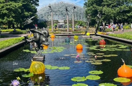 A serene pond with colorful glass orbs and lily pads, surrounded by sculptures and a glass conservatory in the background.