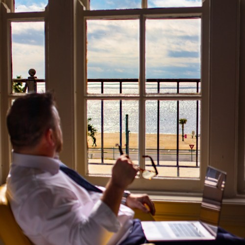 A man in a white shirt sits by a window, gazing at a beach and ocean view, with palm trees in the distance.
