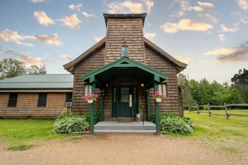 A rustic wooden house with a green entrance, surrounded by greenery and a gravel path, under a cloudy sky.
