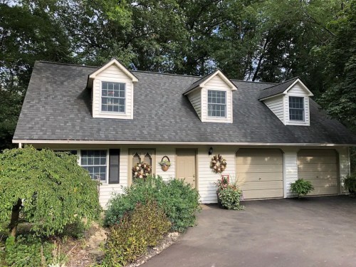 A charming house with a steep roof, two dormer windows, and a landscaped front yard, featuring wreaths on the doors.
