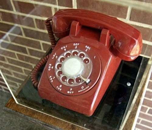 A vintage red rotary phone displayed in a glass case against a brick wall.