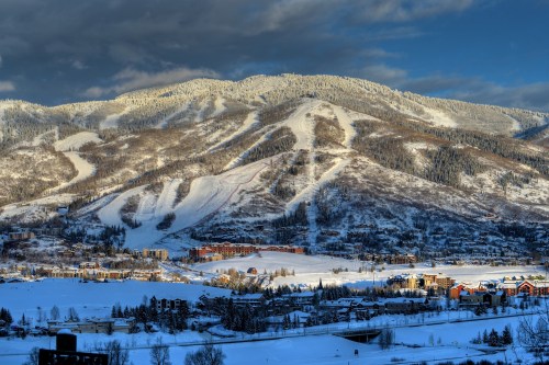 Snow-covered mountains with ski slopes, a valley of trees, and a small town under a partly cloudy sky.