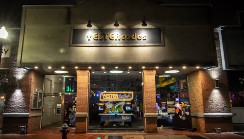A brightly lit arcade entrance at night, featuring the name "MESTERcade" above the door and arcade games visible inside.