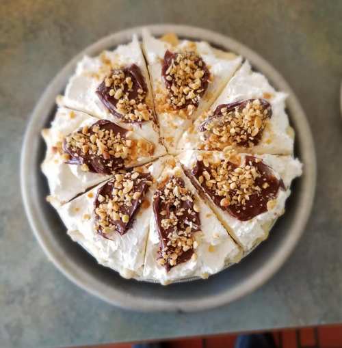 A round cake topped with whipped cream, chocolate, and crushed nuts, sliced into eight pieces on a plate.