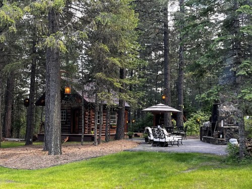 A cozy log cabin surrounded by tall trees, with outdoor seating and a stone fireplace in a serene forest setting.