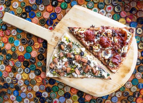 Two slices of pizza on a wooden paddle, surrounded by a colorful array of bottle caps.