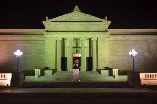 A grand museum building illuminated at night, featuring a statue in front and decorative lamps on either side.