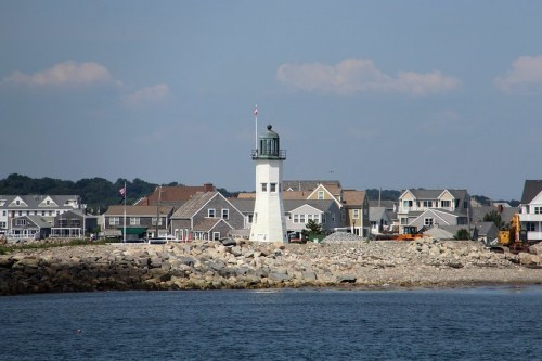 A white lighthouse stands on a rocky shore, surrounded by houses and a calm blue sea under a clear sky.