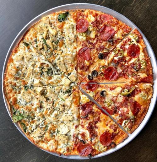 A round pizza divided into two halves: one with white sauce, spinach, and chicken; the other with pepperoni, olives, and green peppers.