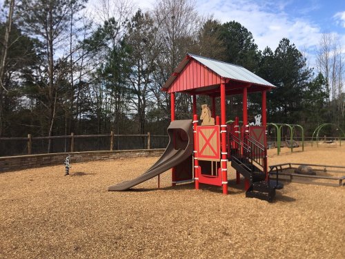 Colorful playground structure with a slide, surrounded by gravel and trees under a blue sky.