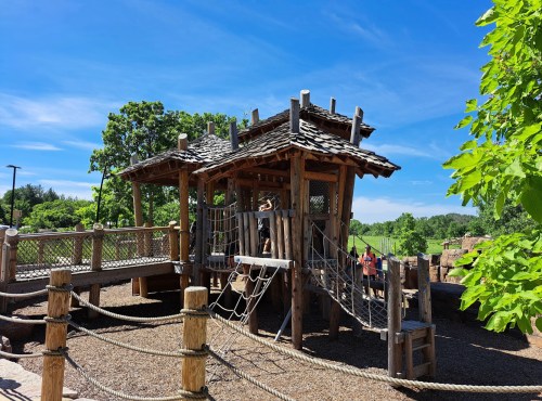 A wooden playground structure with a slide and climbing features, surrounded by greenery and a clear blue sky.