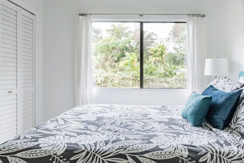 A bright bedroom with a floral-patterned bedspread, white curtains, and a window overlooking greenery.