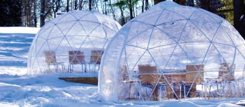 Two transparent igloos with wooden tables and chairs, surrounded by snow and trees in a winter landscape.