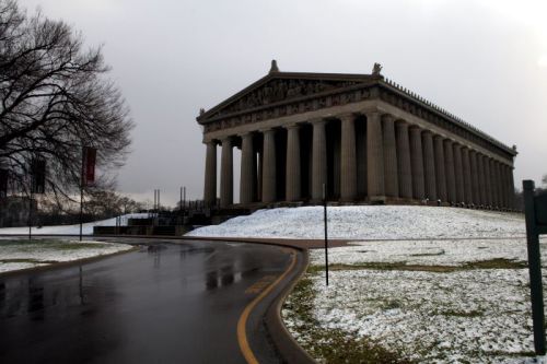 A large classical building with columns, set on a grassy hill, surrounded by a light dusting of snow and a cloudy sky.