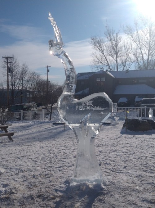 An ice sculpture of a bird stands in a snowy outdoor setting, with trees and buildings in the background.