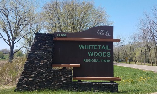 Sign for Whitetail Woods Regional Park, featuring stone base and green lettering, surrounded by trees and grass.
