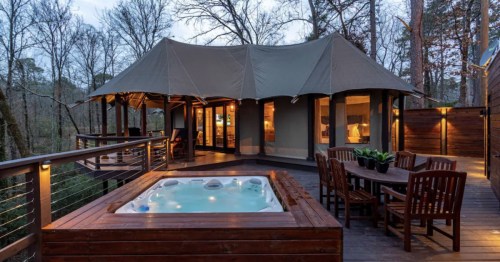 A cozy tent-style cabin with a hot tub on a deck, surrounded by trees in a serene outdoor setting.