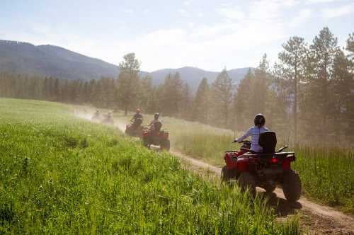 A group of people riding ATVs on a dirt path through a grassy field, surrounded by trees and mountains.