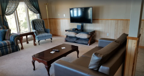 Cozy living room with a TV, two armchairs, a coffee table, and a sofa, featuring wooden paneling and large windows.