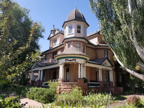 A large, historic Victorian house with a turret, surrounded by greenery and trees under a clear blue sky.