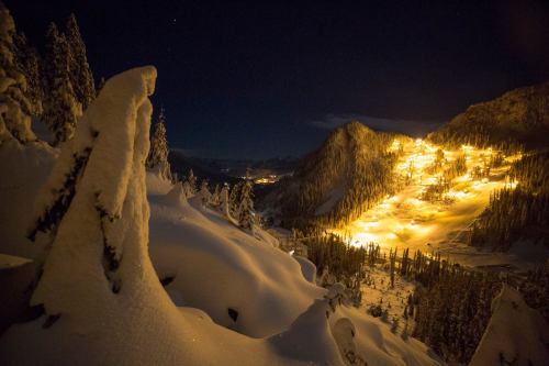 A snowy landscape at night, illuminated by lights from a ski resort nestled in the mountains.