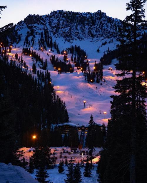 A snowy mountain landscape at dusk, with ski lifts and lights illuminating the slopes and surrounding trees.